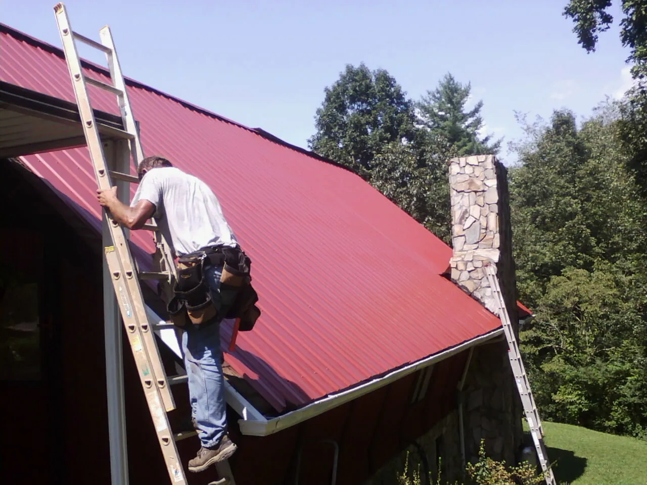 Roofer on a ladder beside a red metal roof and stone chimney — Soffit & Fascia Repair by Roofing Brothers in Saint Marys